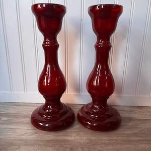 May include: A pair of matching deep red glass candle holders featuring a smooth, polished finish. Each piece has a wide, circular base, a slender, curved stem, and a flared top opening designed to securely hold a standard taper candle.