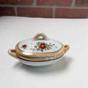 May include: A small, lidded ceramic dish with a floral design. The dish is white with a gold rim and handle. The lid features a cluster of red and white flowers with green leaves. The dish has small handles on either side.