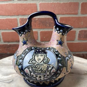 May include: A ceramic vase with a handle and two spouts. The vase features a portrait of a person in a blue and white oval, surrounded by floral and star designs. The vase has a blue handle and base, with pink and tan accents.