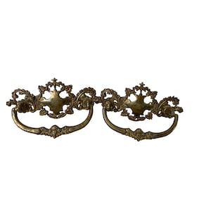 May include: Two antique brass drawer pulls with ornate floral and scrollwork detailing. Each pull features a curved handle and a central decorative element. The brass has a warm, aged patina, suggesting vintage charm.