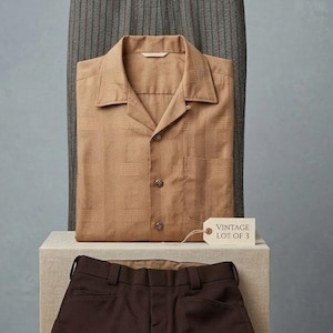 May include: A vintage clothing set featuring a tan button-down shirt, brown trousers, and gray striped pants. The shirt has a textured pattern and the tag reads "VINTAGE LOT OF 3". The items are displayed on a neutral background.