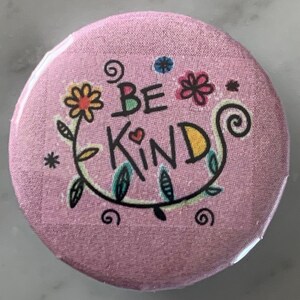 May include: Pink button with a floral design and the text "Be Kind" in black.