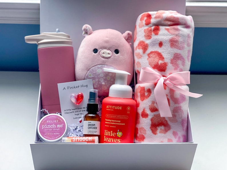 May include: A pink and coral gift box set includes a pink water bottle, a plush pink pig, a pink and coral leopard print throw blanket, a foaming hand soap, a small bottle of essential oil, lip balm, and a small tin of balm. The box also contains a card that says 'A Pocket Hug' and includes a small heart-shaped trinket. The overall theme is self-care and relaxation.