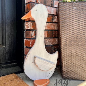 May include: A flat, wooden cutout of a duck featuring a distressed white finish on the body and head, with a bright orange beak and a matching orange foot. The duck has a simple, rounded wing detail and a small, dark dot for an eye.
