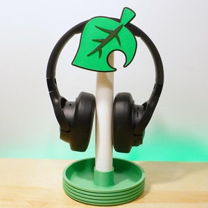 May include: A green leaf-shaped stand with a white pole holds a pair of black headphones. The stand is on a wooden surface.