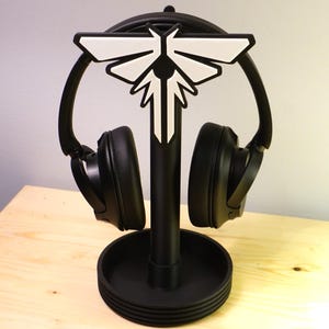 Peut inclure: Casque audio noir posé sur un support noir orné d'un motif blanc et noir. Le support, avec une base circulaire, maintient le casque. Les écouteurs sont positionnés pour mettre en valeur les oreillettes. Le support est en plastique.