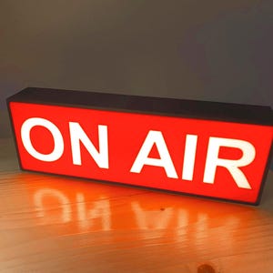 May include: A red rectangular sign with a black frame and white lettering that reads "ON AIR".