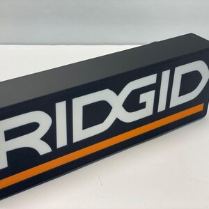 Puede incluir: Objeto rectangular negro con la palabra "RIDGID" en letras blancas grandes. Una franja naranja recorre la parte inferior del texto. Un cable USB es visible en el fondo.