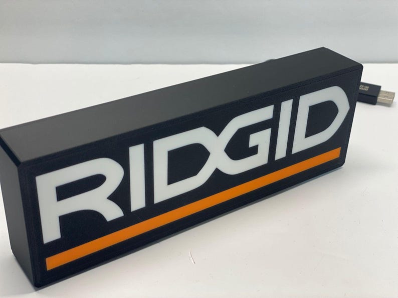 Puede incluir: Objeto rectangular negro con la palabra "RIDGID" en letras blancas grandes. Una franja naranja recorre la parte inferior del texto. Un cable USB es visible en el fondo.