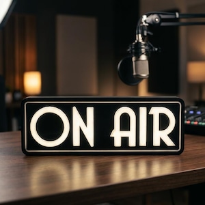 Peut inclure: Un panneau rectangulaire noir avec des lettres blanches lumineuses indiquant ON AIR. Le panneau présente une police de caractères nette et audacieuse avec une fine bordure blanche entourant le texte, créant un affichage contrasté.