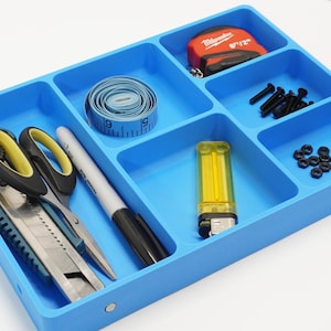 May include: A bright blue rectangular organiser tray with six compartments containing various tools and hardware. Items include a utility knife, scissors, a marker, a coiled measuring tape, a Milwaukee tape measure, black screws, and small nuts.