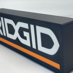 Puede incluir: Letrero rectangular negro con la palabra "RIDGID" en letras blancas grandes. Una franja naranja recorre la parte inferior. El letrero est&aacute; enchufado con un cable negro.