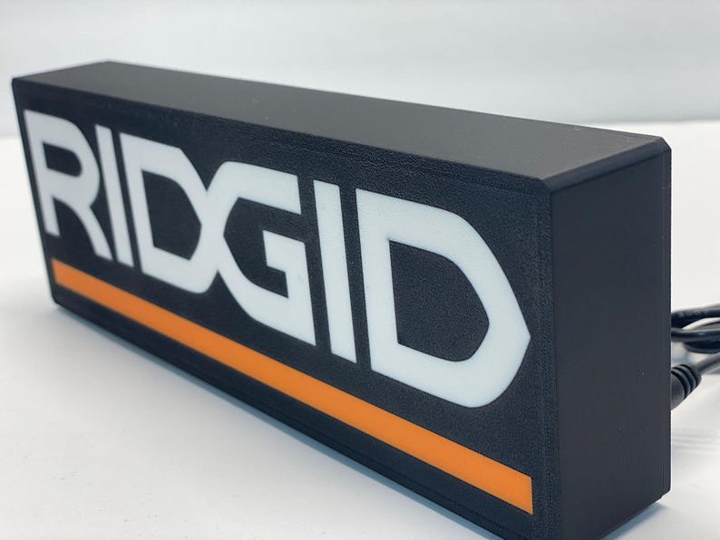 Puede incluir: Letrero rectangular negro con la palabra "RIDGID" en letras blancas grandes. Una franja naranja recorre la parte inferior. El letrero est&aacute; enchufado con un cable negro.