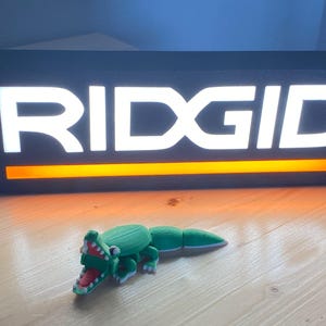 Puede incluir: Un juguete de cocodrilo articulado verde y blanco con boca roja est&aacute; frente a un letrero negro con el texto blanco "RIDGID" y una franja naranja. El letrero est&aacute; iluminado.