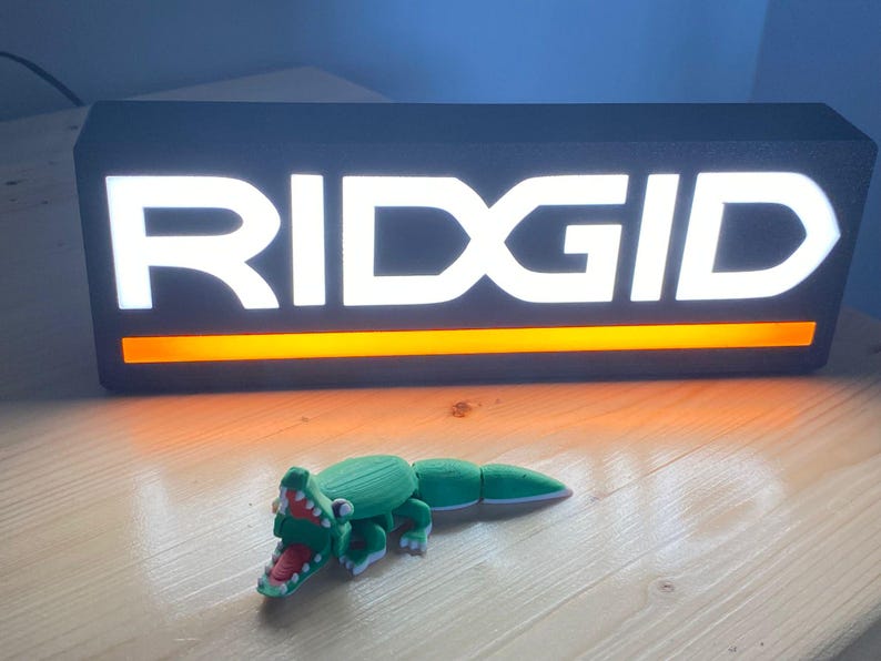 Puede incluir: Un juguete de cocodrilo articulado verde y blanco con boca roja est&aacute; frente a un letrero negro con el texto blanco "RIDGID" y una franja naranja. El letrero est&aacute; iluminado.