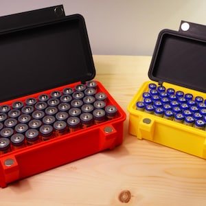 May include: Two open, rectangular cases, one red and one yellow, each filled with small cylindrical objects. The red case contains silver-topped objects, while the yellow case holds blue-topped ones. Both cases have black lids.