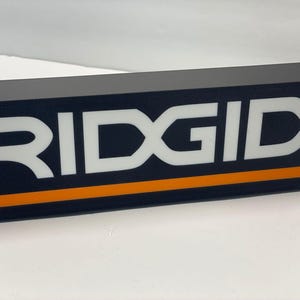 Puede incluir: Letrero rectangular negro con la palabra "RIDGID" en letras blancas grandes. Una franja naranja recorre la parte inferior. El letrero es probablemente para una marca de herramientas o equipos.