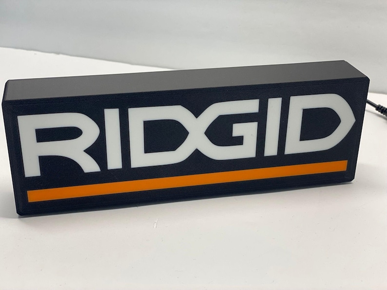 Puede incluir: Letrero rectangular negro con la palabra "RIDGID" en letras blancas grandes. Una franja naranja recorre la parte inferior. El letrero es probablemente para una marca de herramientas o equipos.
