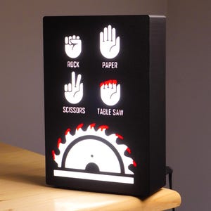 May include: Black rectangular light box with white illuminated graphics of rock, paper, scissors, and table saw. The table saw graphic has red accents. The text labels are in white.
