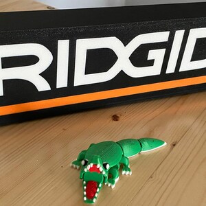 Puede incluir: Un letrero rectangular negro con la palabra "RIDGID" en blanco y una franja naranja. Un juguete de cocodrilo verde impreso en 3D con boca roja est&aacute; frente al letrero. Los objetos est&aacute;n sobre una superficie de madera.