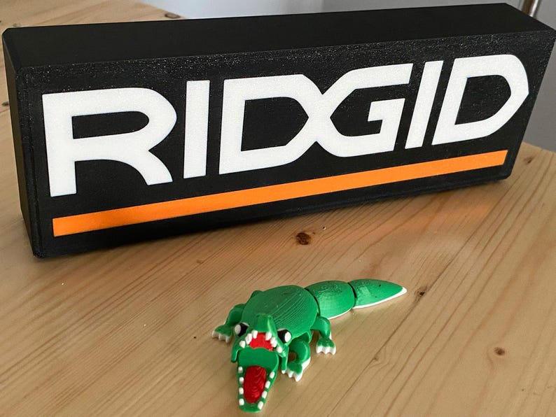 Puede incluir: Un letrero rectangular negro con la palabra "RIDGID" en blanco y una franja naranja. Un juguete de cocodrilo verde impreso en 3D con boca roja est&aacute; frente al letrero. Los objetos est&aacute;n sobre una superficie de madera.