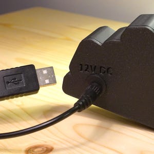 Puede incluir: Un dispositivo electr&oacute;nico negro con un cable USB conectado. El dispositivo tiene el texto "12V DC" impreso. El cable USB est&aacute; conectado a un puerto USB.
