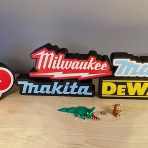 Puede incluir: Una colecci&oacute;n de letreros decorativos con los logotipos de las marcas de herramientas el&eacute;ctricas Milwaukee, Makita y DeWalt. Tambi&eacute;n incluye un dise&ntilde;o de nube roja y peque&ntilde;as figuras de juguete de un cocodrilo y un mono. Los letreros son negros con detalles en blanco, rojo y amarillo.