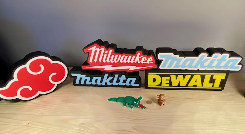 Puede incluir: Una colecci&oacute;n de letreros decorativos con los logotipos de las marcas de herramientas el&eacute;ctricas Milwaukee, Makita y DeWalt. Tambi&eacute;n incluye un dise&ntilde;o de nube roja y peque&ntilde;as figuras de juguete de un cocodrilo y un mono. Los letreros son negros con detalles en blanco, rojo y amarillo.