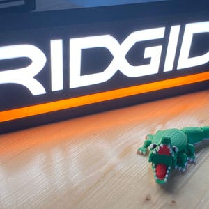 Puede incluir: Un letrero iluminado negro y naranja con la palabra "RIDGID" en blanco. Una figura de cocodrilo impresa en 3D, verde y roja, est&aacute; frente al letrero. El letrero y la figura est&aacute;n sobre una superficie de madera.