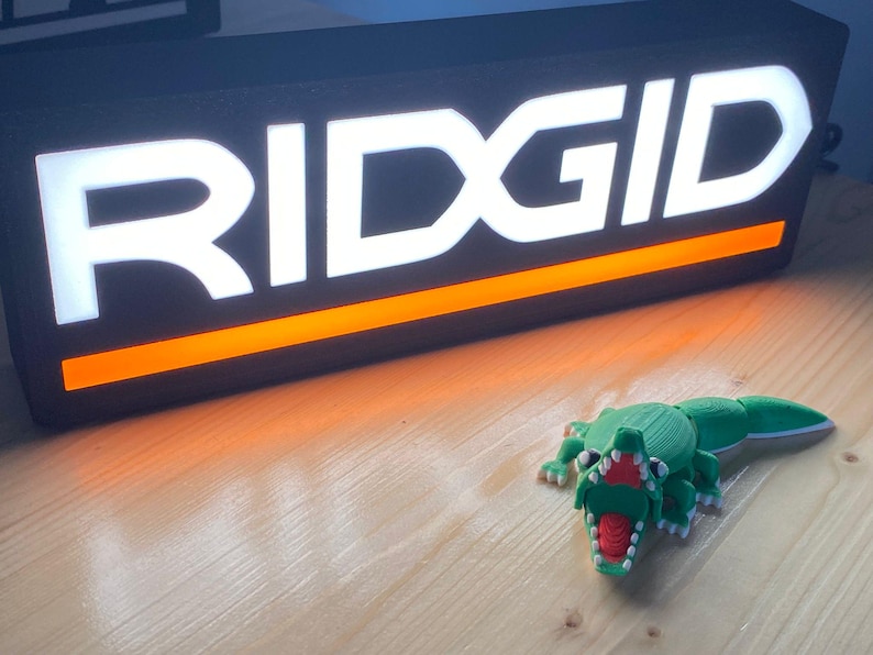 Puede incluir: Un letrero iluminado negro y naranja con la palabra "RIDGID" en blanco. Una figura de cocodrilo impresa en 3D, verde y roja, est&aacute; frente al letrero. El letrero y la figura est&aacute;n sobre una superficie de madera.