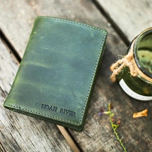 Op de afbeelding: Een groene leren portemonnee met de tekst "NOAH RIVER" in reliëf wordt getoond op een houten oppervlak. Een glazen pot met touwdetail staat in de buurt. De portemonnee heeft een gestikte rand.