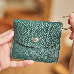 May include: A small, dark green leather wallet with a textured surface. The wallet has a flap closure secured by a silver snap and a silver key ring attached. The wallet is held in a person's hands.
