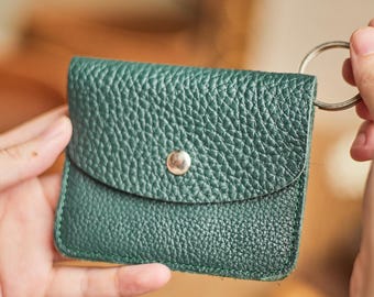 May include: A small, dark green leather wallet with a textured surface. The wallet has a flap closure secured by a silver snap and a silver key ring attached. The wallet is held in a person's hands.