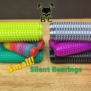 May include: A collection of cylindrical, spiky textured rollers arranged in two stacks. The left stack features neon yellow, rainbow-gradient, and green rollers. The right stack includes metallic silver, purple, and black rollers. Text reads Small! Silent Bearings.