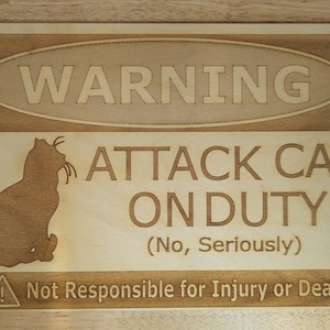 May include: A rectangular light-colored wooden sign featuring a laser-engraved cat silhouette and the text WARNING ATTACK CAT ON DUTY (No, Seriously) and Not Responsible for Injury or Death with a small warning triangle icon in the bottom corner.