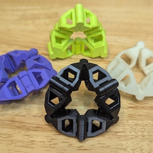 May include: An assortment of 3D-printed geometric shapes in various colors, including purple, green, black, and white. Each shape is composed of interlocking segments, forming a circular design. The objects are displayed on a wooden surface.