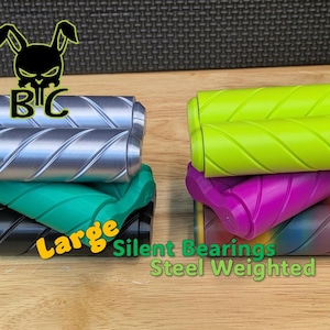 May include: A collection of cylindrical fidget rollers with diagonal grooves, featuring silver, lime green, teal, black, purple, and rainbow colors. Text on the image includes a black rabbit skull logo with the letters BC, and the words Large, Silent Bearings, and Steel Weighted.