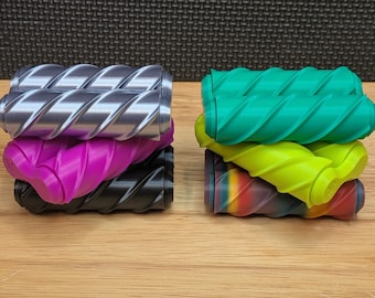 May include: Two stacks of colorful, cylindrical objects with a twisted design. One stack features silver, magenta, and black. The other stack includes green, yellow, and a rainbow-colored piece. The objects are on a wooden surface.