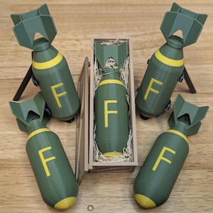 May include: A collection of novelty items shaped like bombs, in olive green with yellow accents and the letter "F". One is in a wooden box with packing material, and the others are on small stands. The background is a wooden surface.