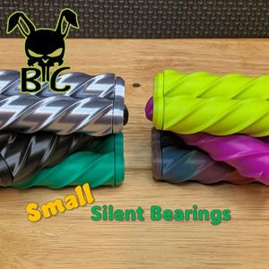 May include: Two stacks of cylindrical silent bearings with a spiral grooved texture. The left stack includes silver, black, and green units. The right stack features neon yellow, purple, and blue units. Text reads BC with a rabbit logo and Small Silent Bearings.