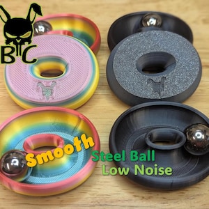 May include: Four circular fidget toys with a central hole and a steel ball bearing. Two toys feature a rainbow gradient finish, while two are matte black. The text "BC" with a rabbit skull logo, "Smooth", and "Steel Ball Low Noise" are visible.