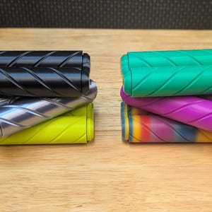 May include: A collection of colorful bicycle handle grips. The grips are in various colors, including black, silver, yellow, green, purple, and a rainbow gradient. Each grip has a textured, diagonal pattern for enhanced grip and control.