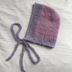 May include: A knitted baby bonnet in shades of lavender and pink. The bonnet features horizontal stripes of pink and lavender, with matching lavender ties for securing. The soft, textured knitwear is displayed on a white background.