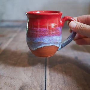 May include: A handcrafted ceramic mug, featuring a striking red glaze that fades into purple and brown tones. The mug has a rounded form and a robust handle. The mug is held by a hand, highlighting its design and size.