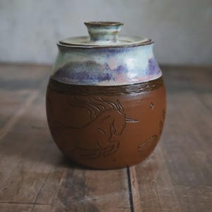 May include: A ceramic jar with a lid. The brown body features an embossed unicorn design. The rim and lid are light blue and purple. The jar sits on a wooden surface.