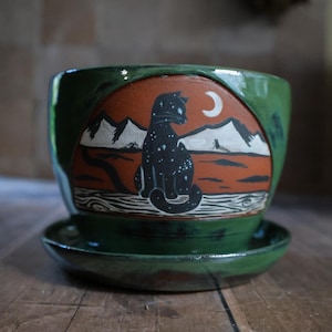 May include: A green ceramic plant pot with a matching saucer. The pot has a hand-painted design of a black cat with white stars, mountains, and a crescent moon. The background is orange and white. Suitable for small plants.