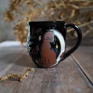 May include: Black ceramic mug with a brown cat silhouette on a white crescent moon. The mug has a black handle and is decorated with leaves, mushrooms, and gold accents. The mug is on a wooden surface.