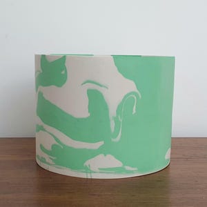 May include: A cylindrical plant pot with a marbled design. The pot features a light green and off-white pattern. The pot is sitting on a wooden surface.