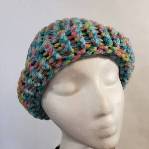 May include: A colourful knitted beanie hat with a teal, pink and yellow colour scheme. The hat has a sparkly finish and is made with a chunky yarn.