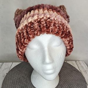 May include: A plush, handmade cat ear beanie in shades of brown, mauve, and cream. The hat is made of a soft, textured material and features two small, upright ears. The beanie is displayed on a white mannequin head.
